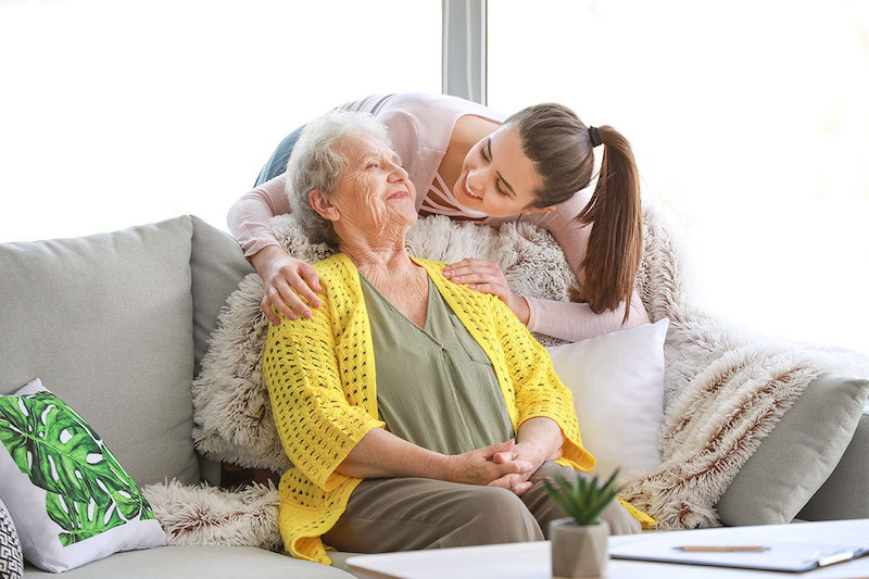 Eine Seniorin im Sessel wird von einer Frau von hinten umarmt