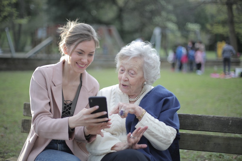 Junge Frau zeigt einer Seniorin was auf dem Handy, während sie auf der Parkbank sitzen